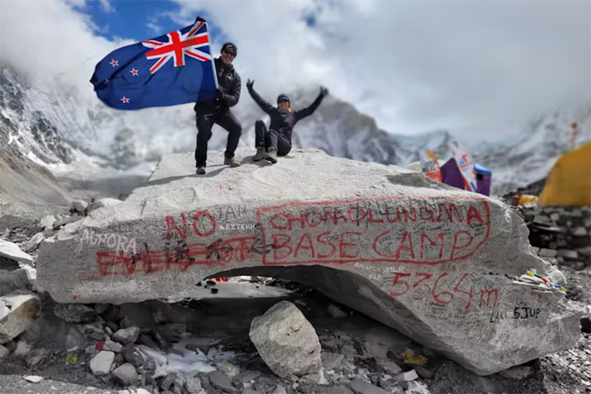 Two people holding the NZ flag atop a large rock with spraypainted words on it. 