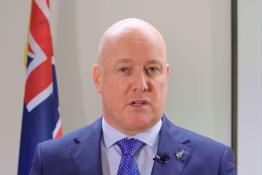 Head and shoulders shot of Prime Minister Chrisotopher Luxon standing next to a NZ flag.
