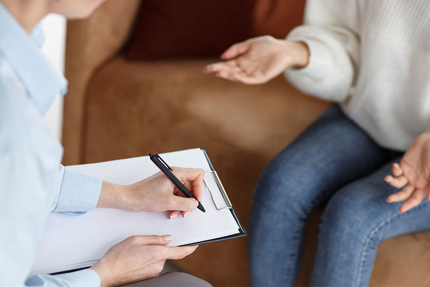 Psychologist Talking With Female Patient Taking Notes During Appointment Sitting In Office. 
